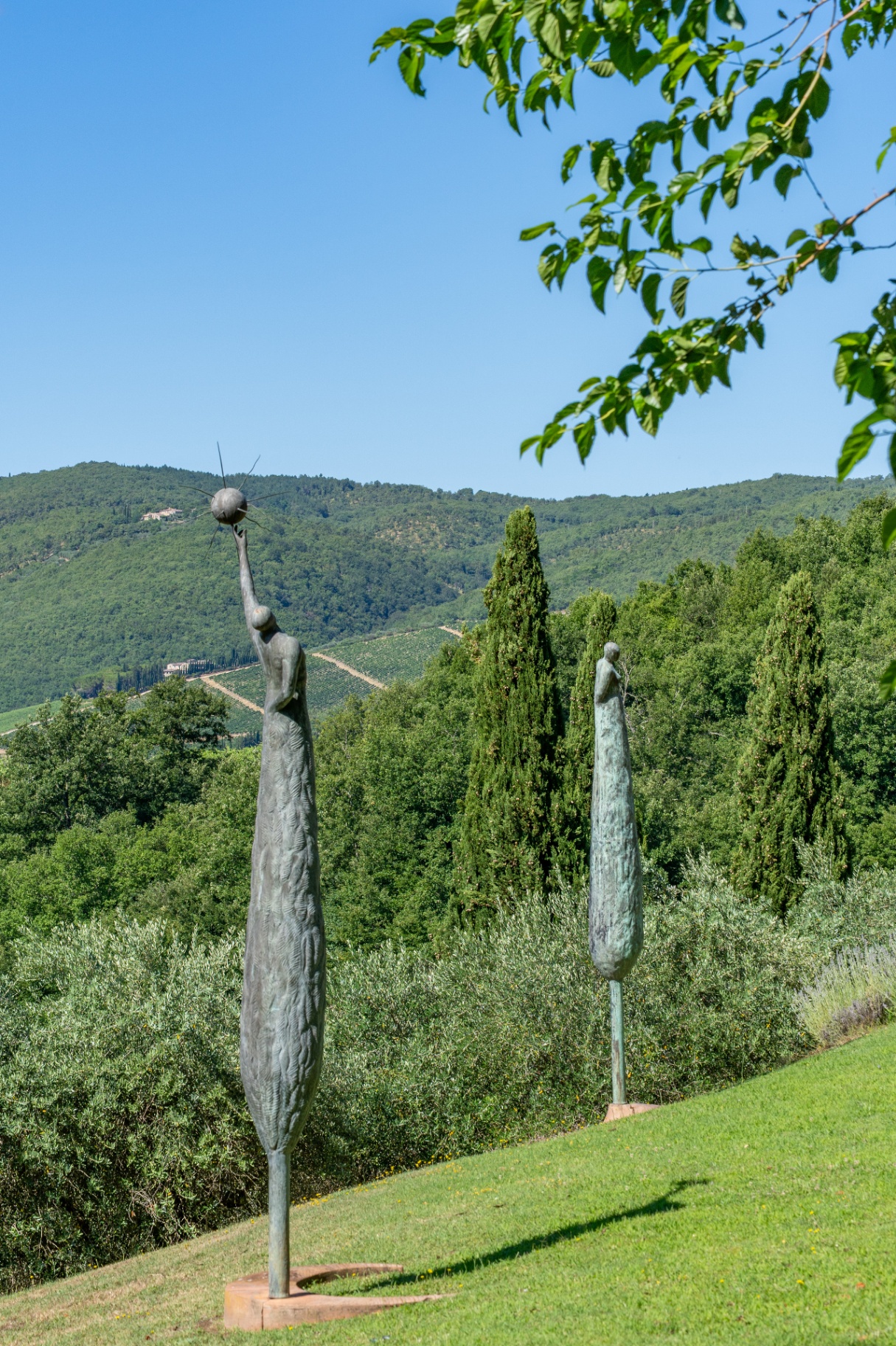 Tall bronze figural sculptures echoing cypress trees against the Chianti hills